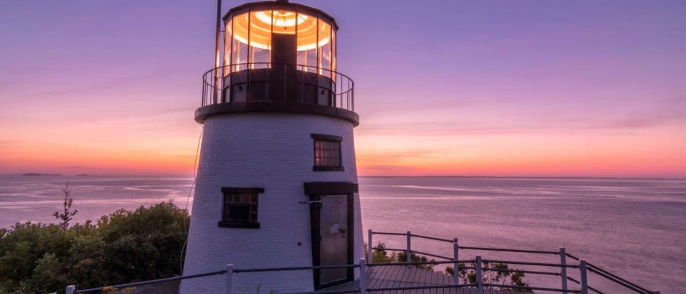 The lighthouse at Owl’s Head is aglow at dawn. The horizon over the ocean is pink and yellow from the rising sun.