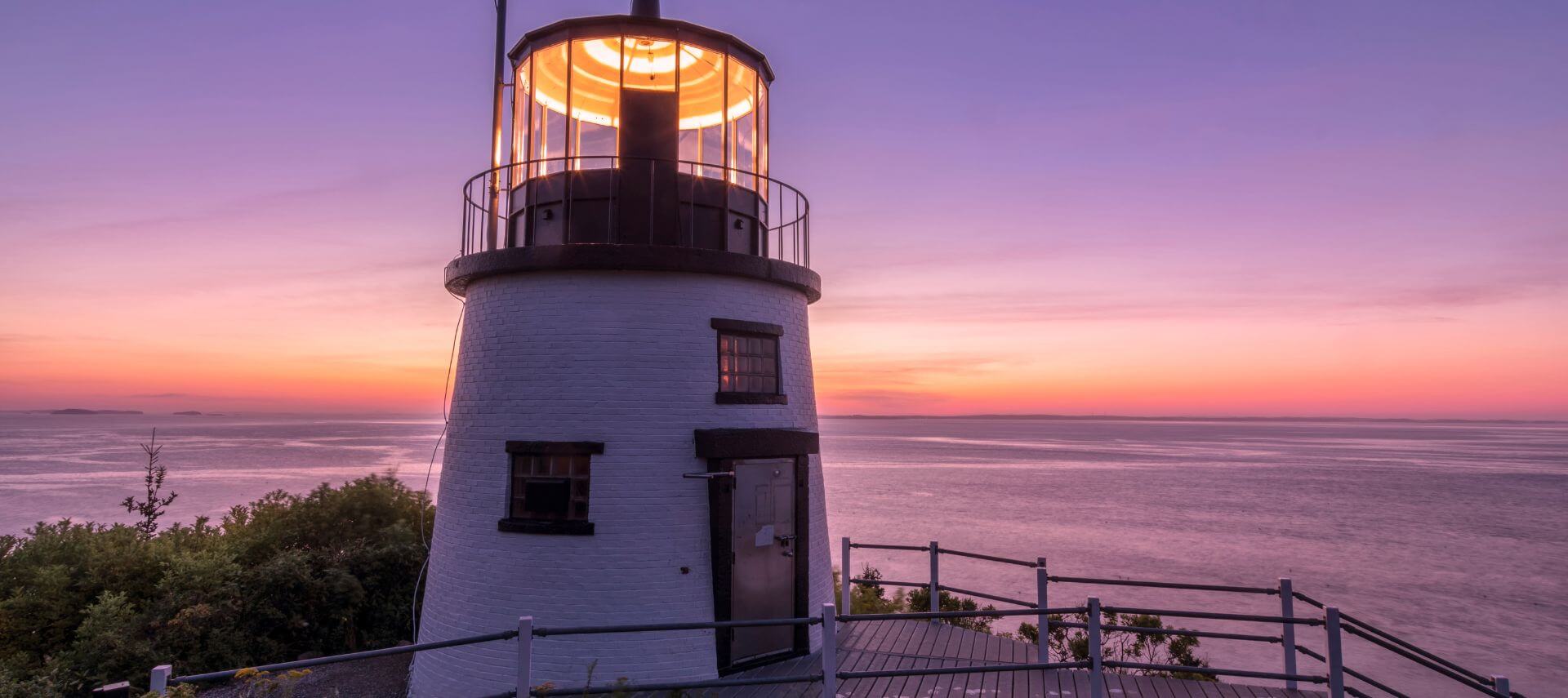 The lighthouse at Owl’s Head is aglow at dawn. The horizon over the ocean is pink and yellow from the rising sun.