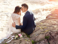 Couple sitting on the rocks at the edge of the ocean on their wedding day with a bouquet set beside them