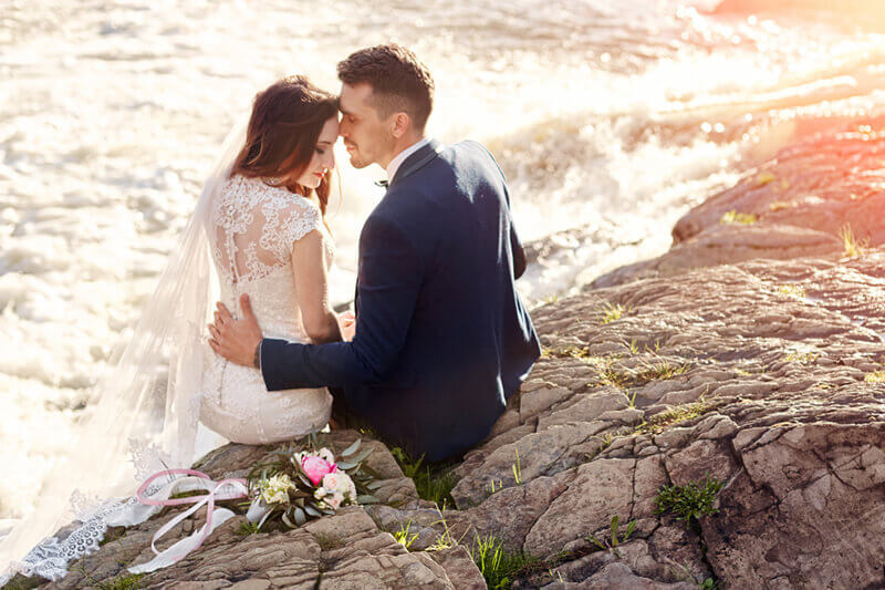 Couple sitting on the rocks at the edge of the ocean on their wedding day with a bouquet set beside them