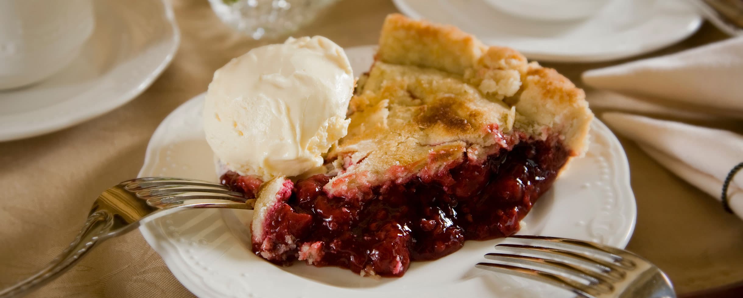 slice of red berry pie with vanilla ice cream a la mode on a white plate with two forks