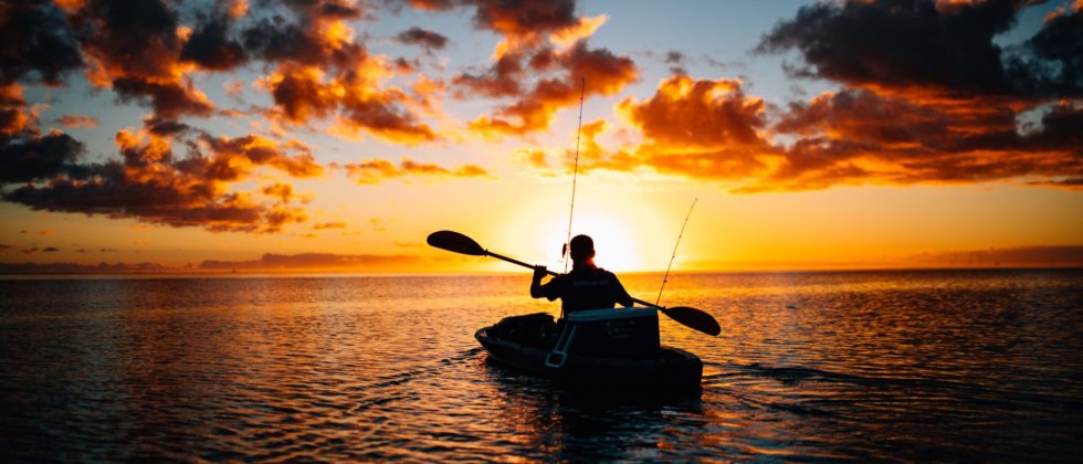 Silhouette of a man kayaking at sunset amidst a brilliant sky of deep gold, orange and red