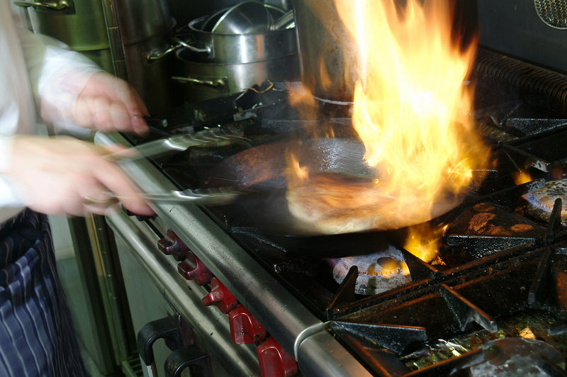 professional stovetop with pan of food being flambed by hand of a chef