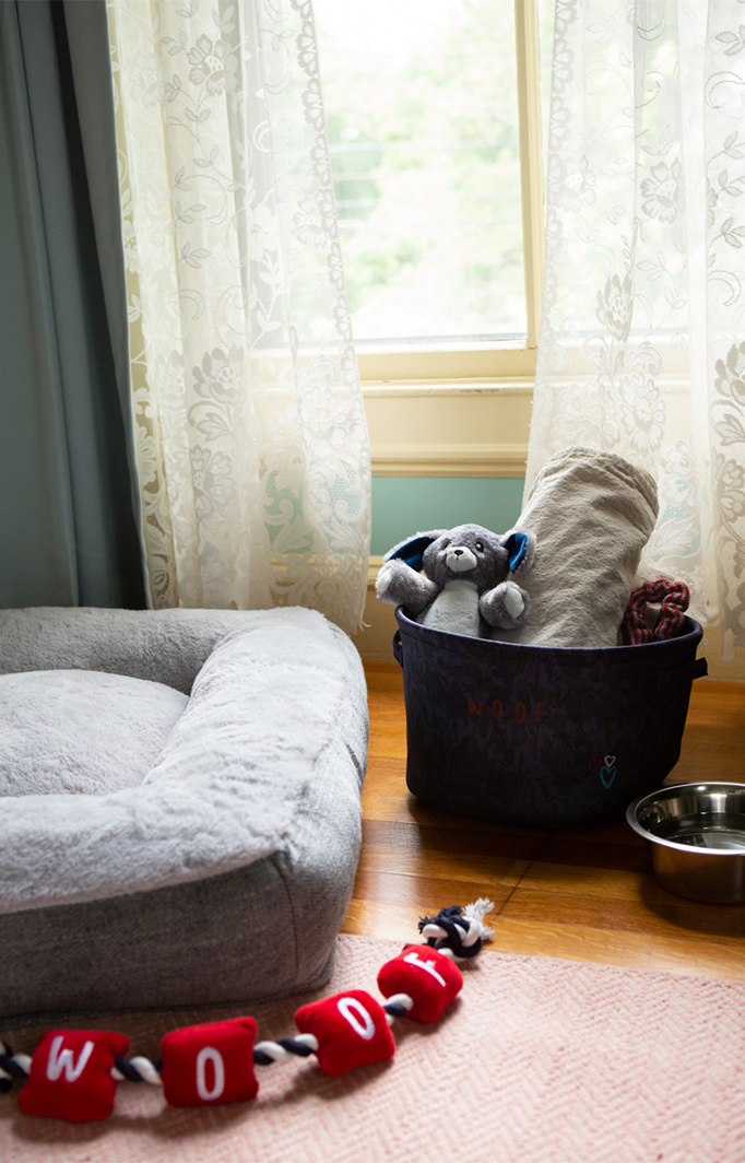 Fluffy grey dog bed laying on a hard wood floor next to a bucket of dog toys