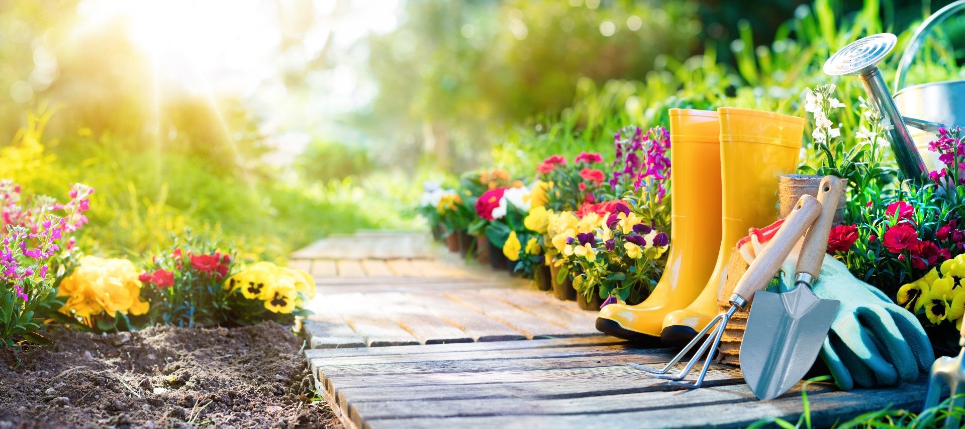 Wooden garden path through a floral garden with boots and garden tools in the forefront