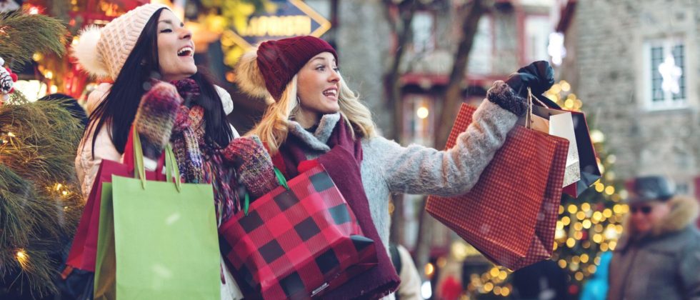 Two women wearing winter hats and holding bags from holiday shopping in a small town.