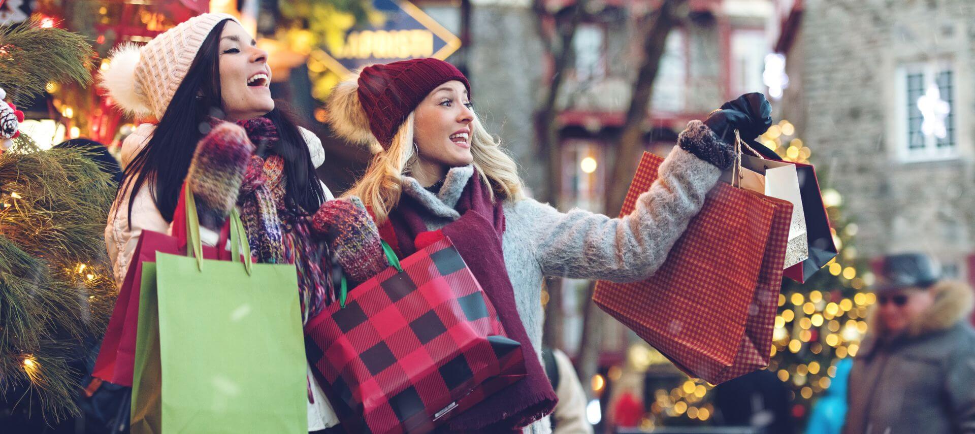 Two women wearing winter hats and holding bags from holiday shopping in a small town.