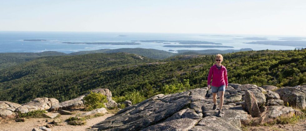 A woman is hiking above the coast of Maine. A view of the Atlantic Ocean in the background.