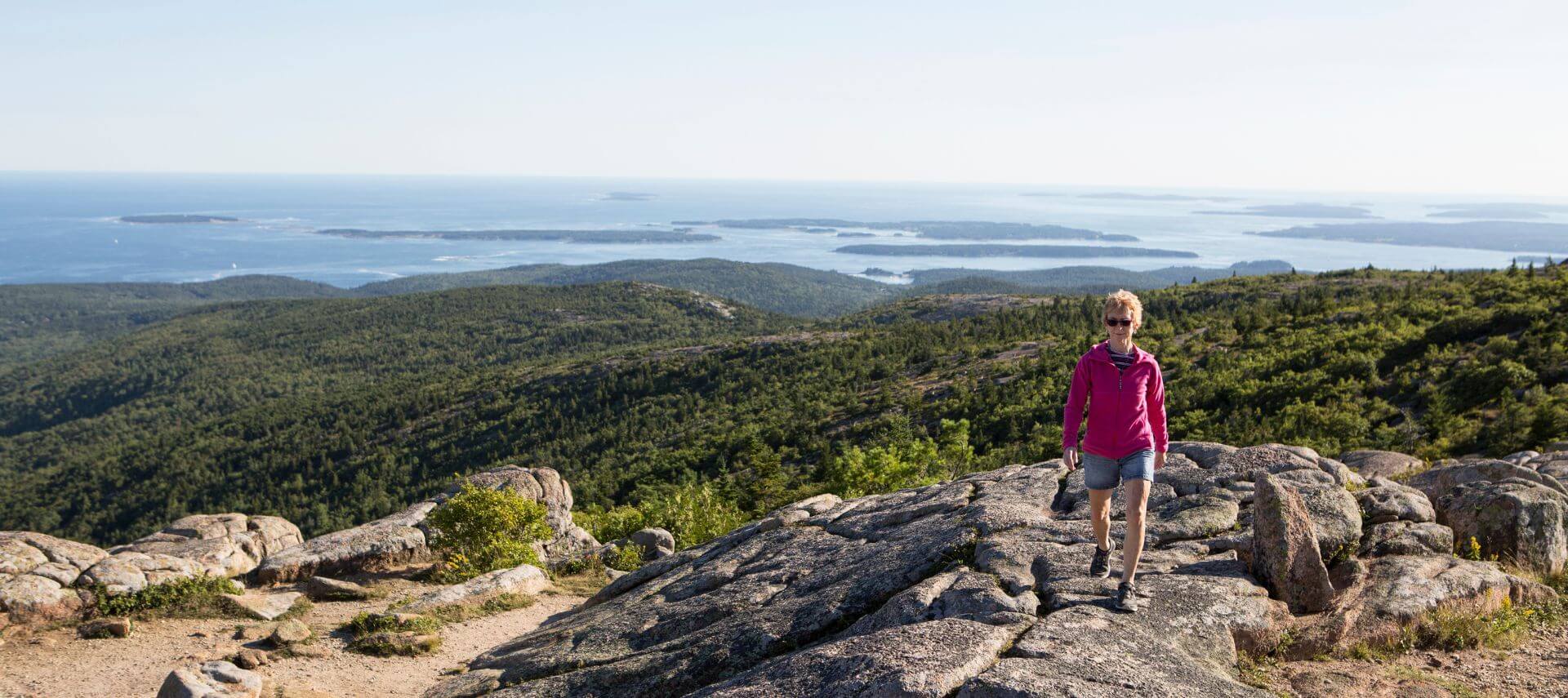 A woman is hiking above the coast of Maine. A view of the Atlantic Ocean in the background.