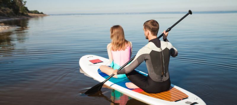 A couple sitting on a SUP wearing wetsuits